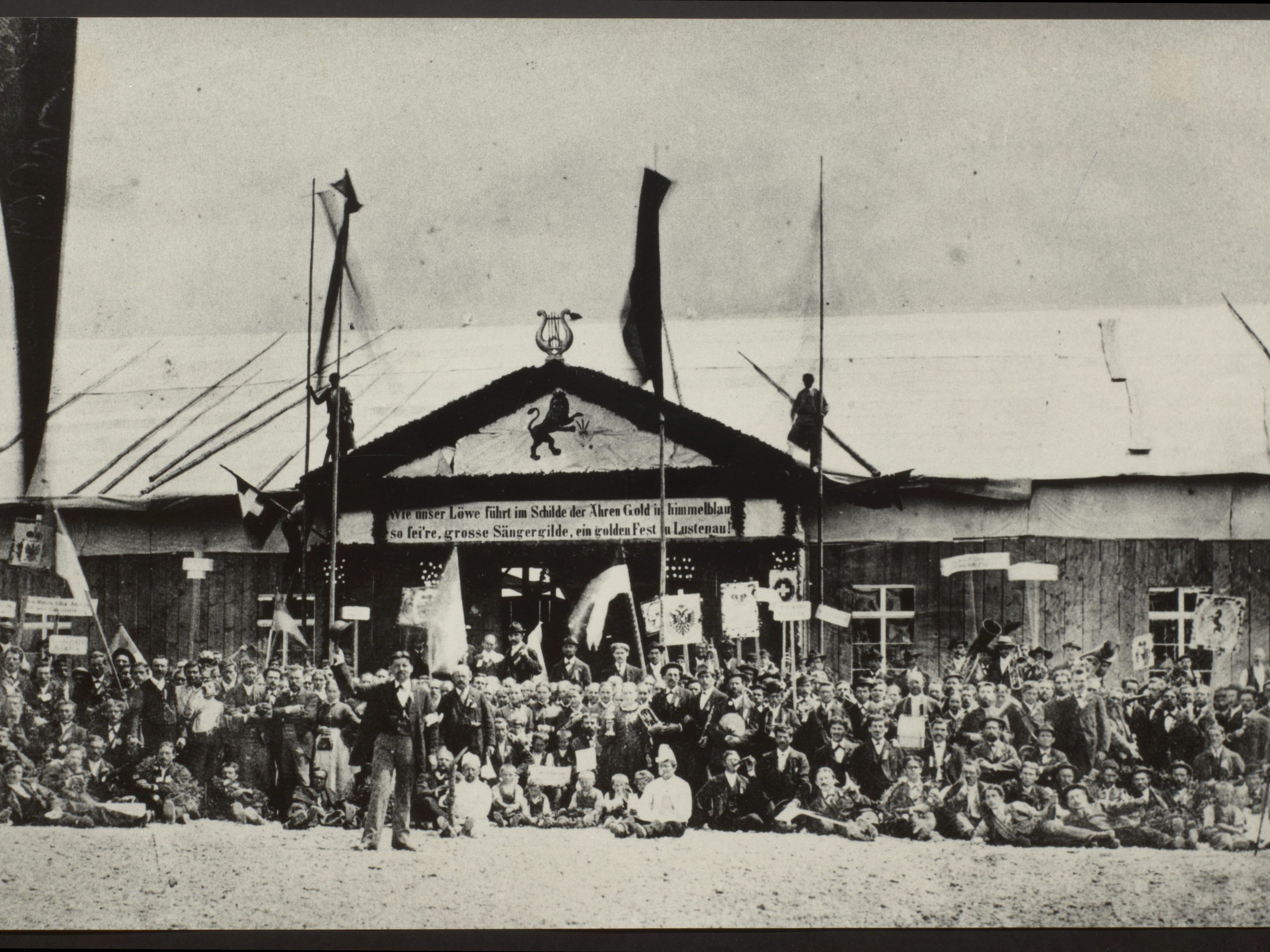 Vermutlich das älteste Foto der Sammlung: Landessängerfest im Jahr 1884 auf dem Platz neben der Volksschule Kirchdorf. Die Tafel am hinteren Gebäude zeigt das Lustenauer Wappen und den Spruch: „Wie unser Löwe führt im Schilde der Ähren Gold in himmelblau / so fei're, grosse Sängergilde, ein golden Fest in Lustenau!". Vermutlich das älteste Foto der Sammlung: Landessängerfest im Jahr 1884 auf dem Platz neben der Volksschule Kirchdorf. Die Tafel am hinteren Gebäude zeigt das Lustenauer Wappen und den Spruch: „Wie unser Löwe führt im Schilde der Ähren Gold in himmelblau / so fei're, grosse Sängergilde, ein golden Fest in Lustenau!".