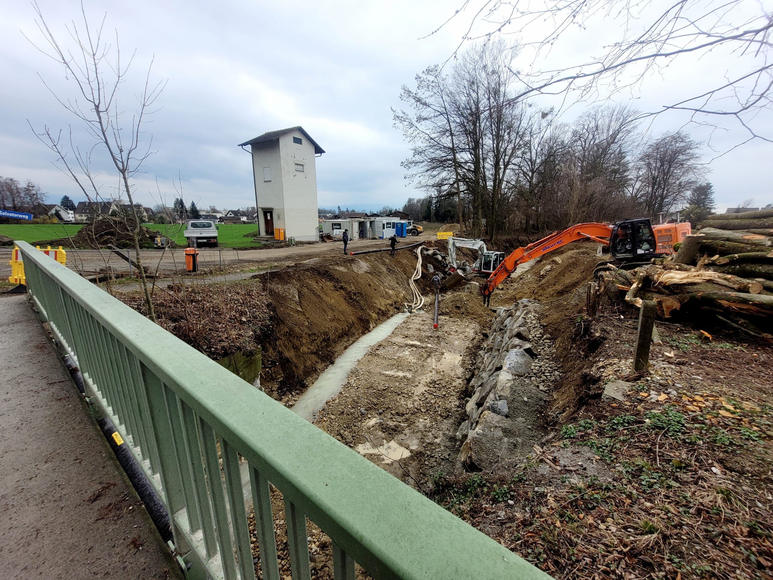 Blick von der Brücke L18 auf die Baustelle Hochwasserschutzprojekt Ruggbach: Prallufersicherung und Nachbettsicherung sowie aktuell die Verlegung der Stromleitung unter dem Ruggbach hindurch.