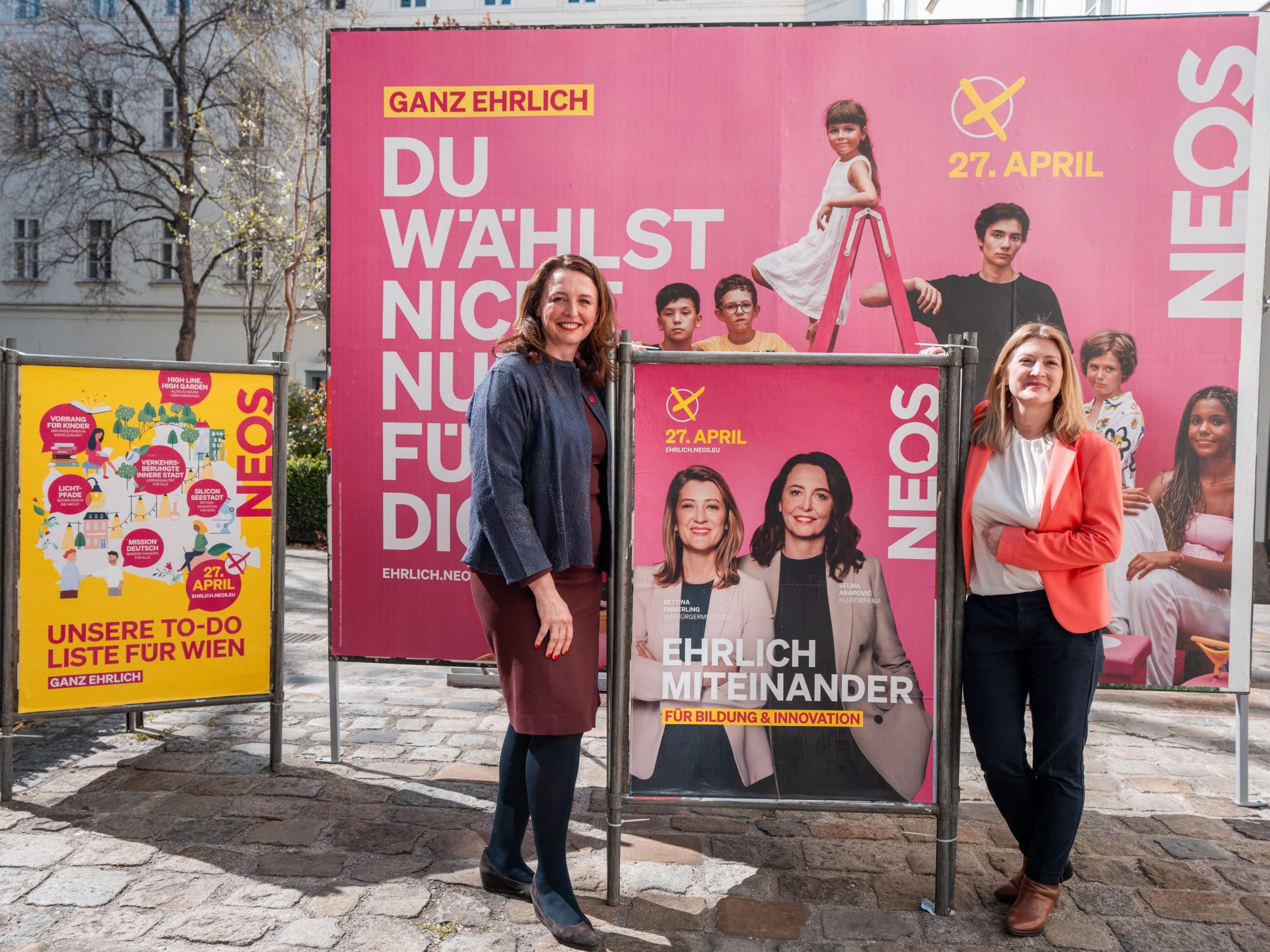 Die NEOS-Spitzenkandidatinnen für die Wien-Wahl neben einem Plakat mit ihnen.