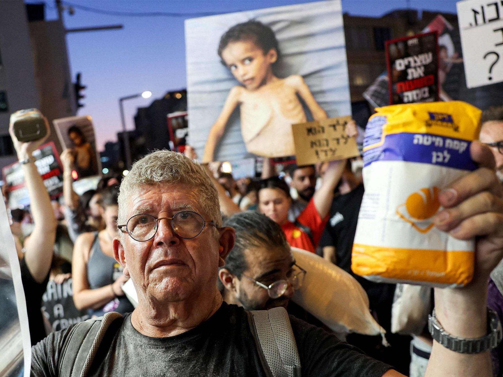 Ein israelischer Aktivist hält einen Sack Mehl und ein Bild eines unter Mangelernährung leidenden Kindes während eines Marsches zum Hauptquartier des israelischen Verteidigungsministeriums in Tel Aviv. Ein israelischer Aktivist hält einen Sack Mehl und ein Bild eines unter Mangelernährung leidenden Kindes während eines Marsches zum Hauptquartier des israelischen Verteidigungsministeriums in Tel Aviv.