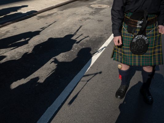 Auf dem Bild sieht man einen Mann, der einen schottischen Kilt trägt. Daneben sieht man die Schatten von mehreren Dudelsack-Spielern.