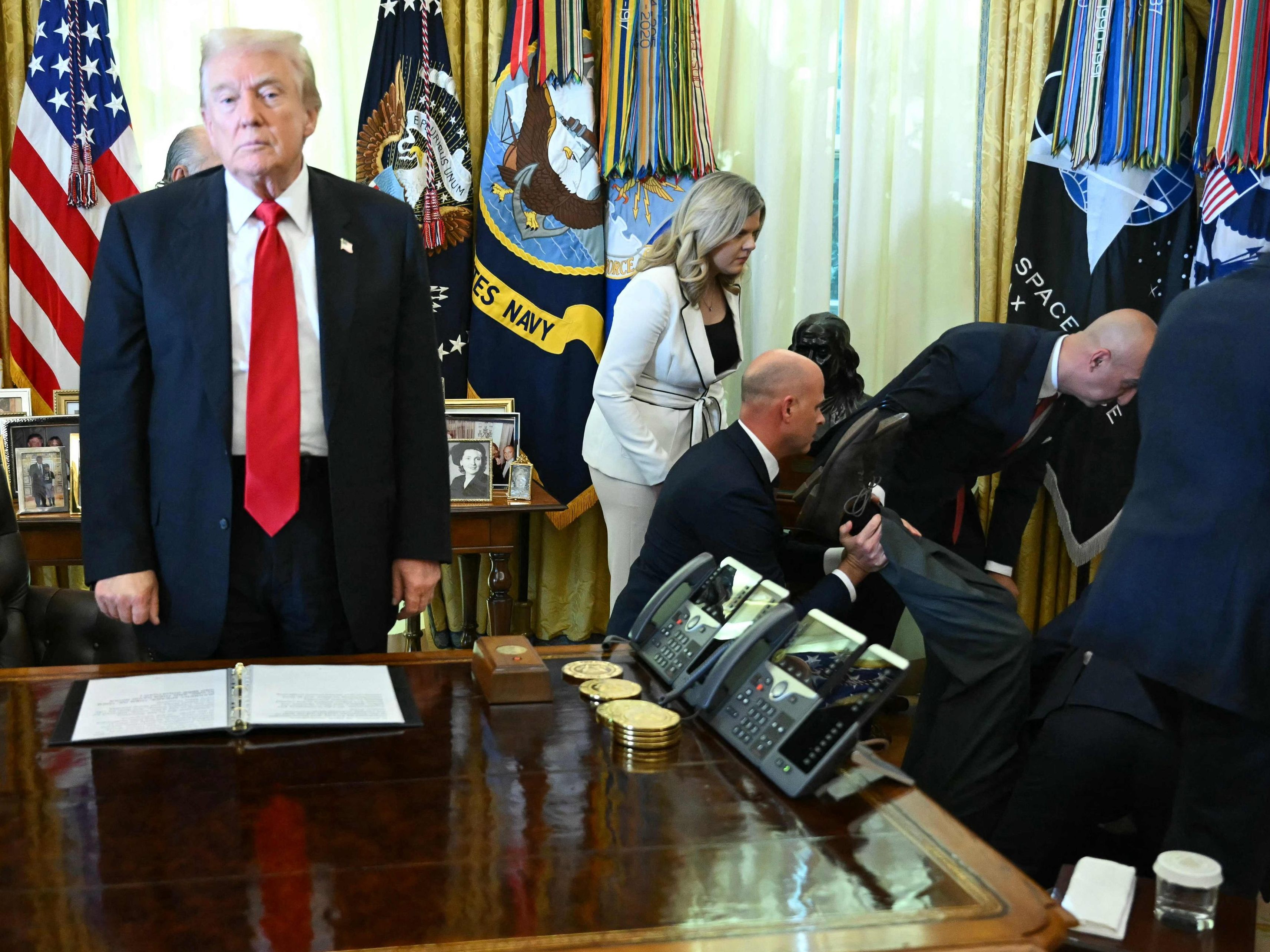 Donald Trump an seinem Schreibtisch im Oval Office, während der kollabierte Mann hinter ihm versorgt wird.